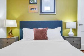 A hotel room with a bed, two bedside tables, a nightstand, a lamp, and a wall with framed artwork. - Hip & Stylish Magazine St. Apt | Balcony & Parking (New Orleans)