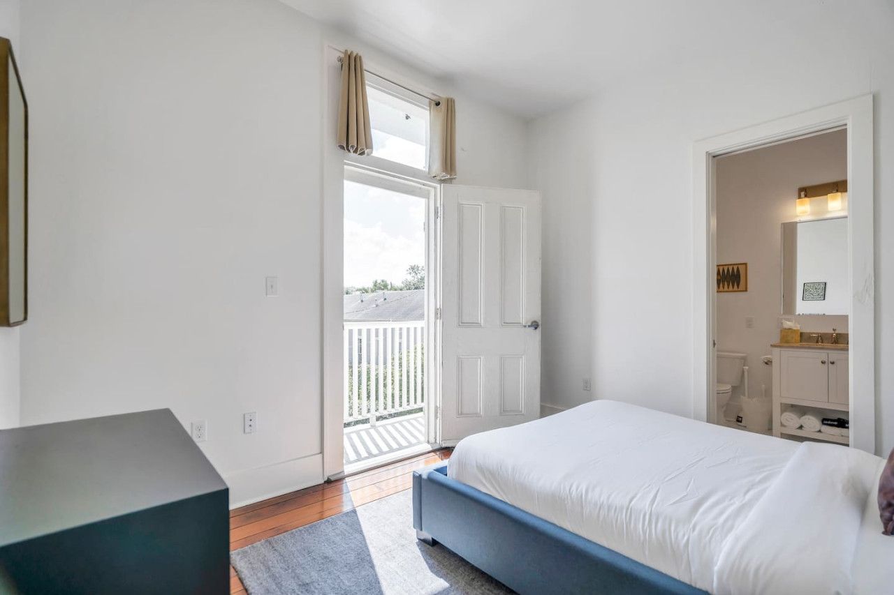 A bedroom with a bed, a bench, a door leading to a balcony, and a bathroom with a sink and mirror.