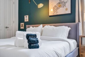 A neatly made bed with white bedding and a stack of towels with 'Makeup' printed on them.