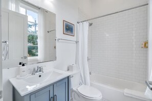 A bathroom with a white sink, mirror, and toilet, a shower with a white curtain, and a window with a view of greenery.