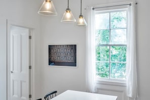 A dining area with a white table, three pendant lights, a framed artwork, and a window with a view of trees.