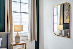 Une pièce avec une grande fenêtre, une lampe sur une petite table et un miroir au mur.