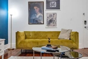 A living room with a mustard yellow sofa, a glass coffee table, and framed artwork on the wall.