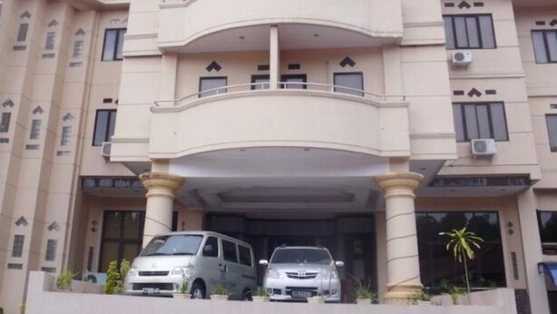 Un edificio de varios pisos con un balcón central, un acceso cubierto y dos coches estacionados.