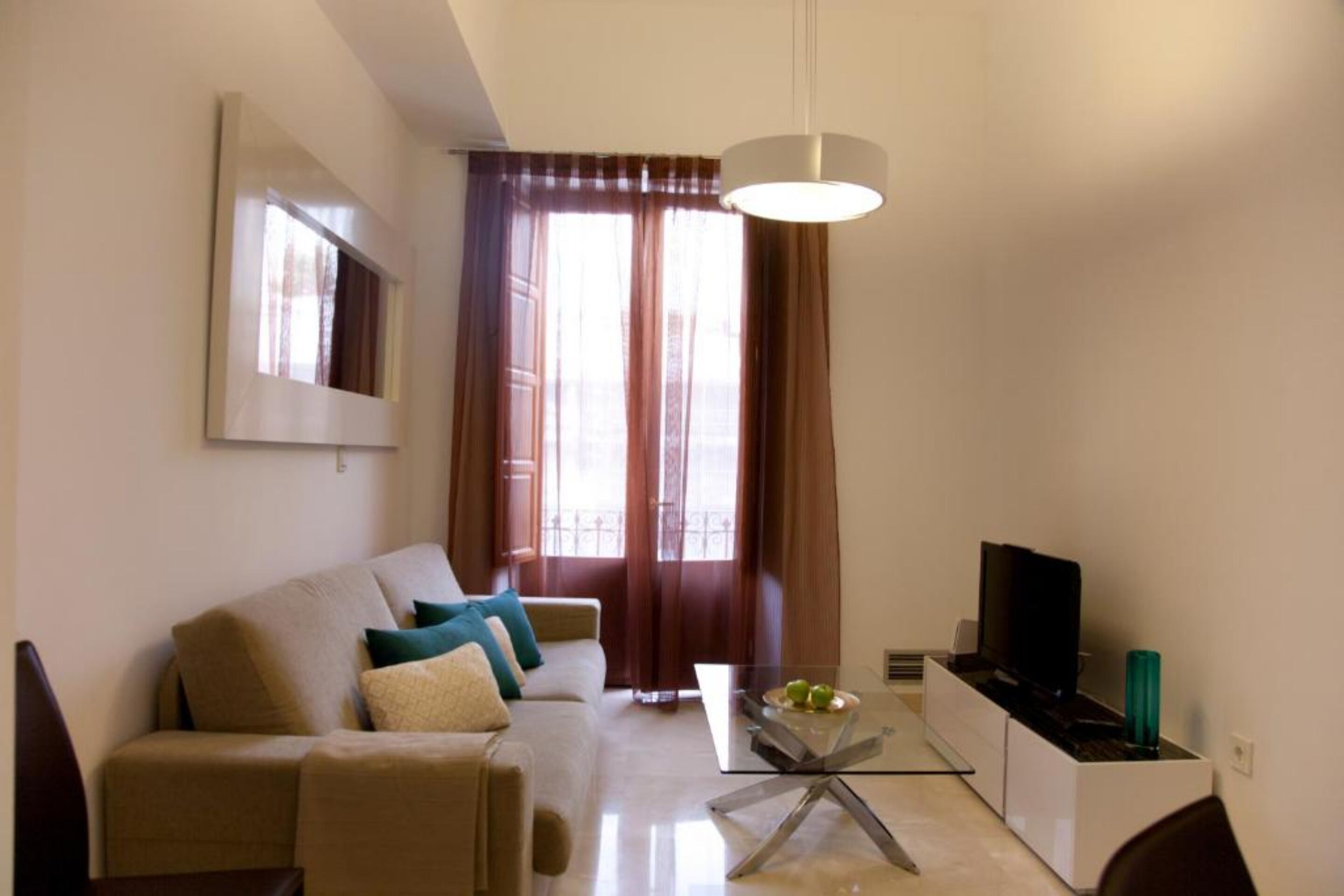 A modern living room with a sofa, a glass coffee table, a TV, and a large window with curtains.