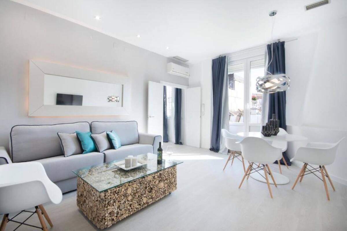 A modern living room with a grey sofa, a glass coffee table, and a dining area with white chairs and a table.