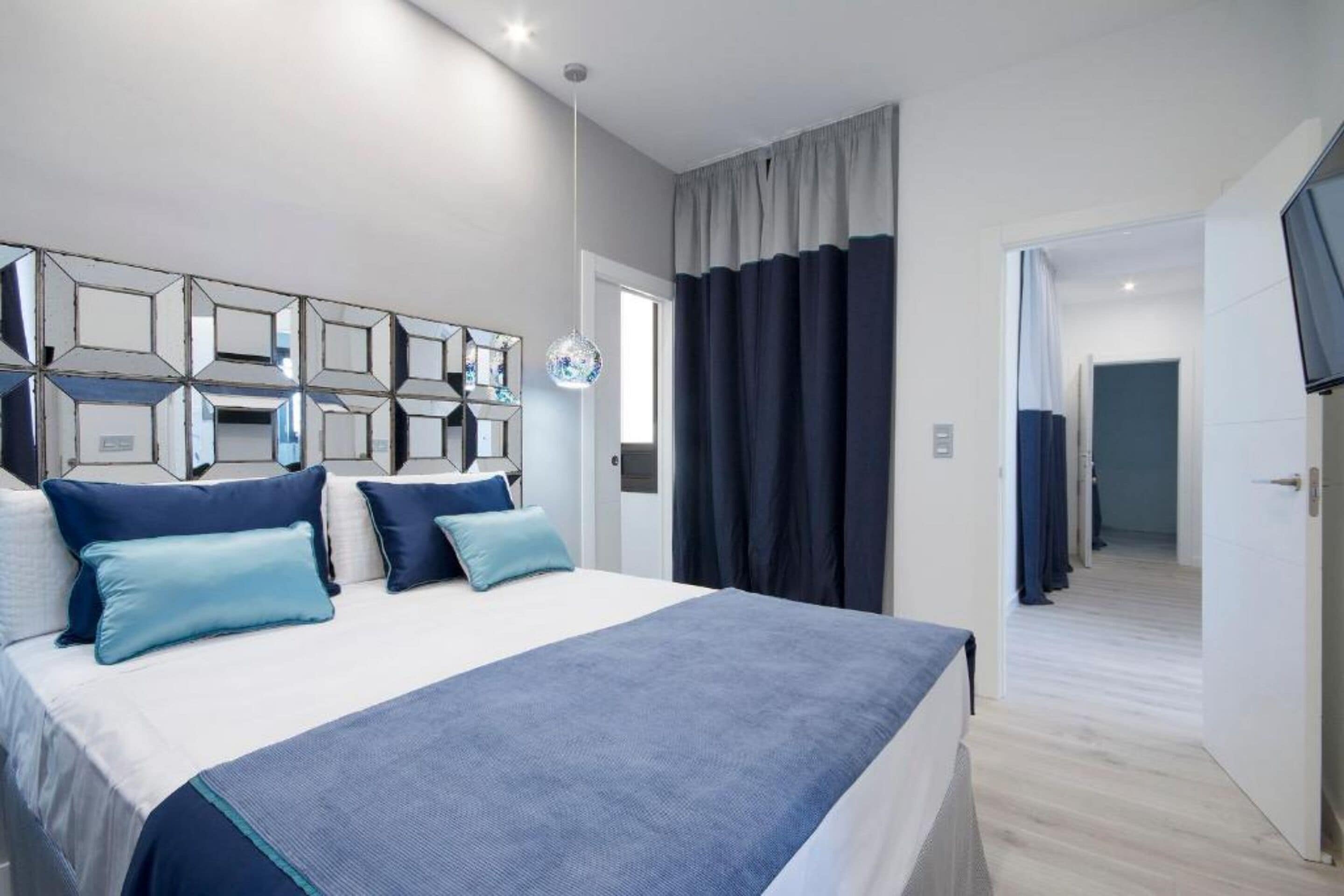 A modern bedroom with a large bed, a mirror with geometric frames, and a view of a hallway.