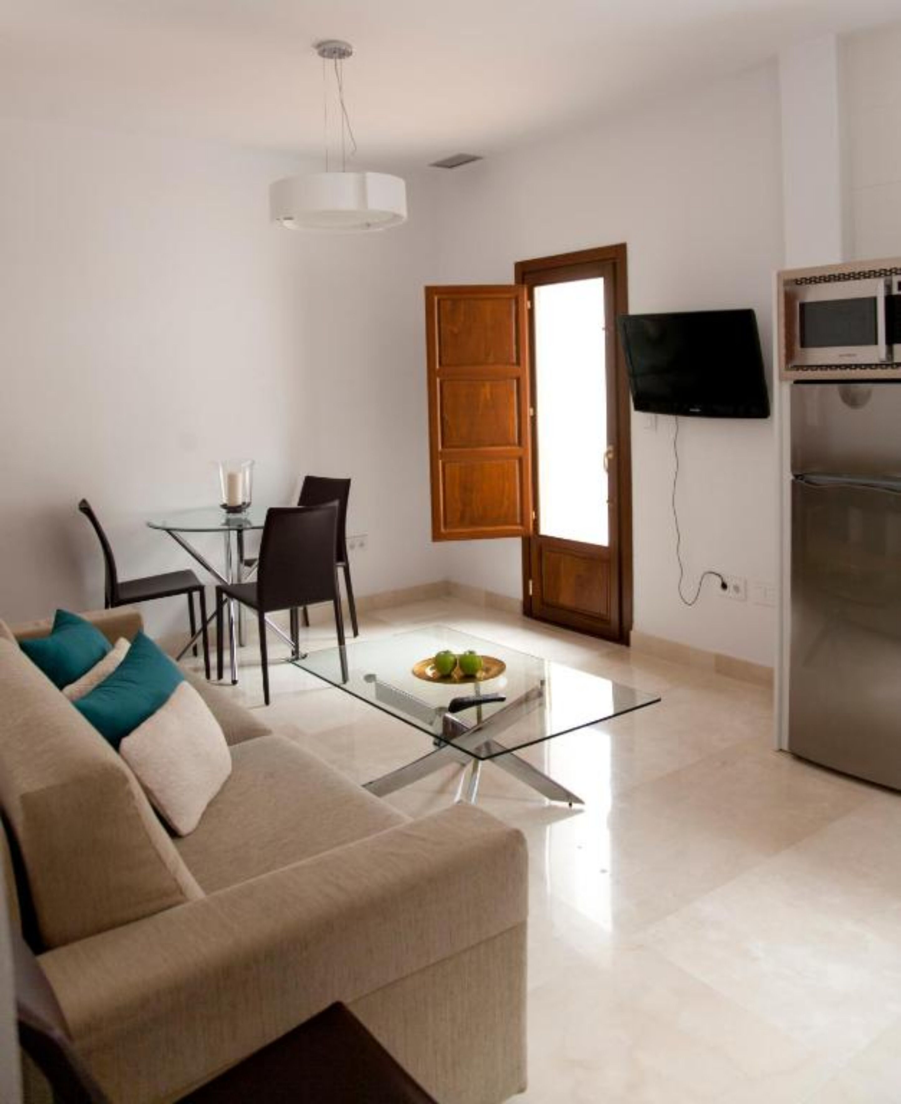 Una sala moderna con un sofá, una mesa de centro de vidrio, una mesa de comedor con sillas, una puerta de madera, un televisor y un refrigerador.