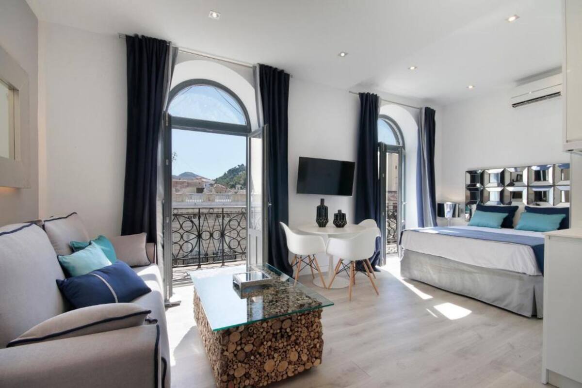 A modern hotel room with a bed, a sofa, a glass coffee table, a TV, and a balcony with a view.