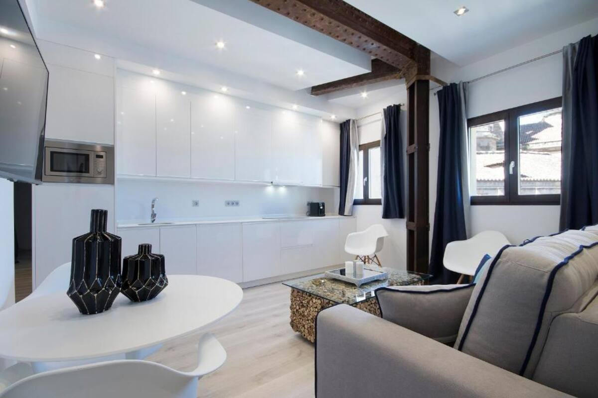 A modern living room with a white sofa, a coffee table, and two black vases. There is a kitchen area with white cabinets and a microwave.