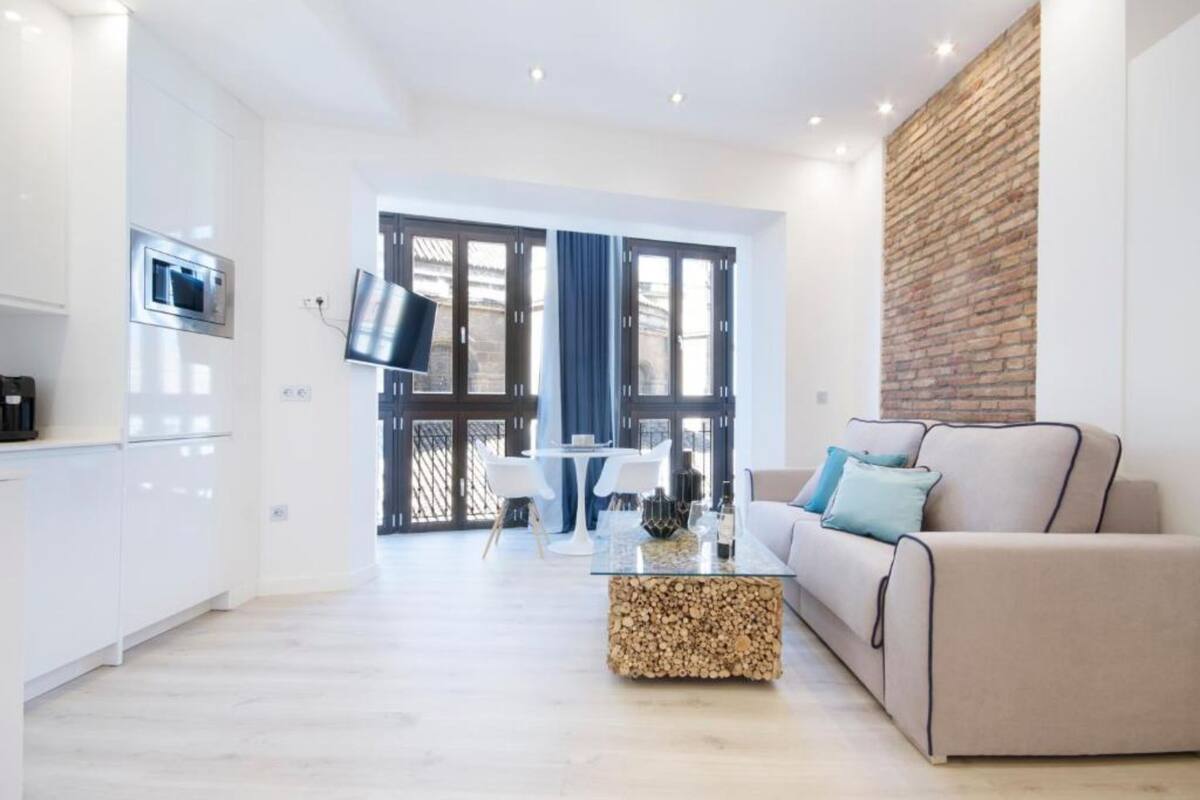 A modern living room with a sofa, a coffee table, and a TV. There is a brick wall and a window with curtains.