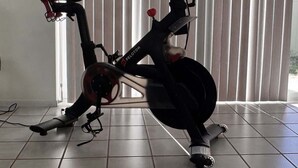 Un vélo d’appartement est installé dans une pièce dont le sol est carrelé et qui possède une fenêtre avec des stores verticaux.