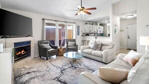 Un salon avec une cheminée, deux fauteuils, un canapé, une table basse en verre et un ventilateur de plafond.