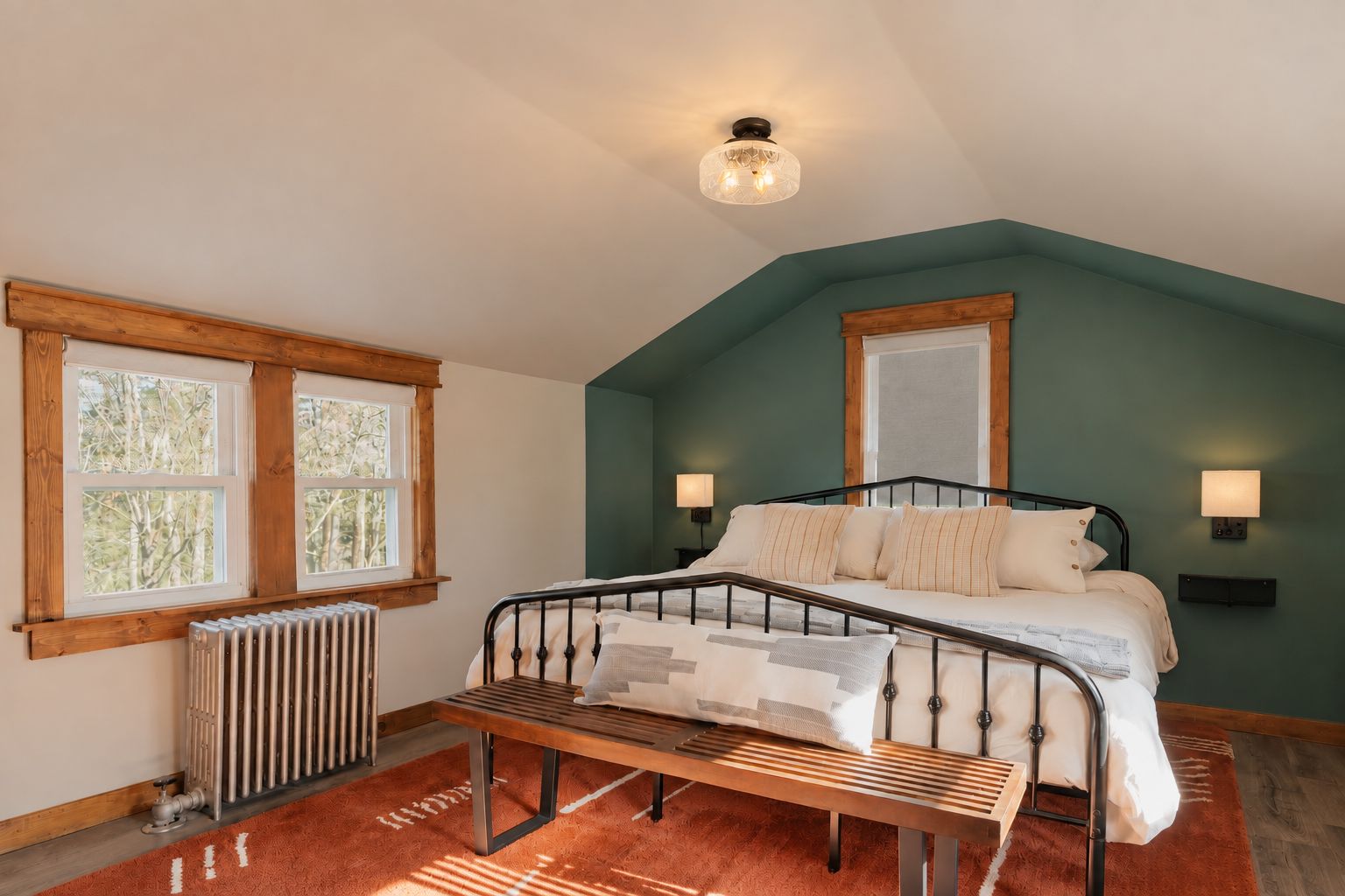 A bedroom with a bed, bedside lamps, a radiator, a window, and a bench.