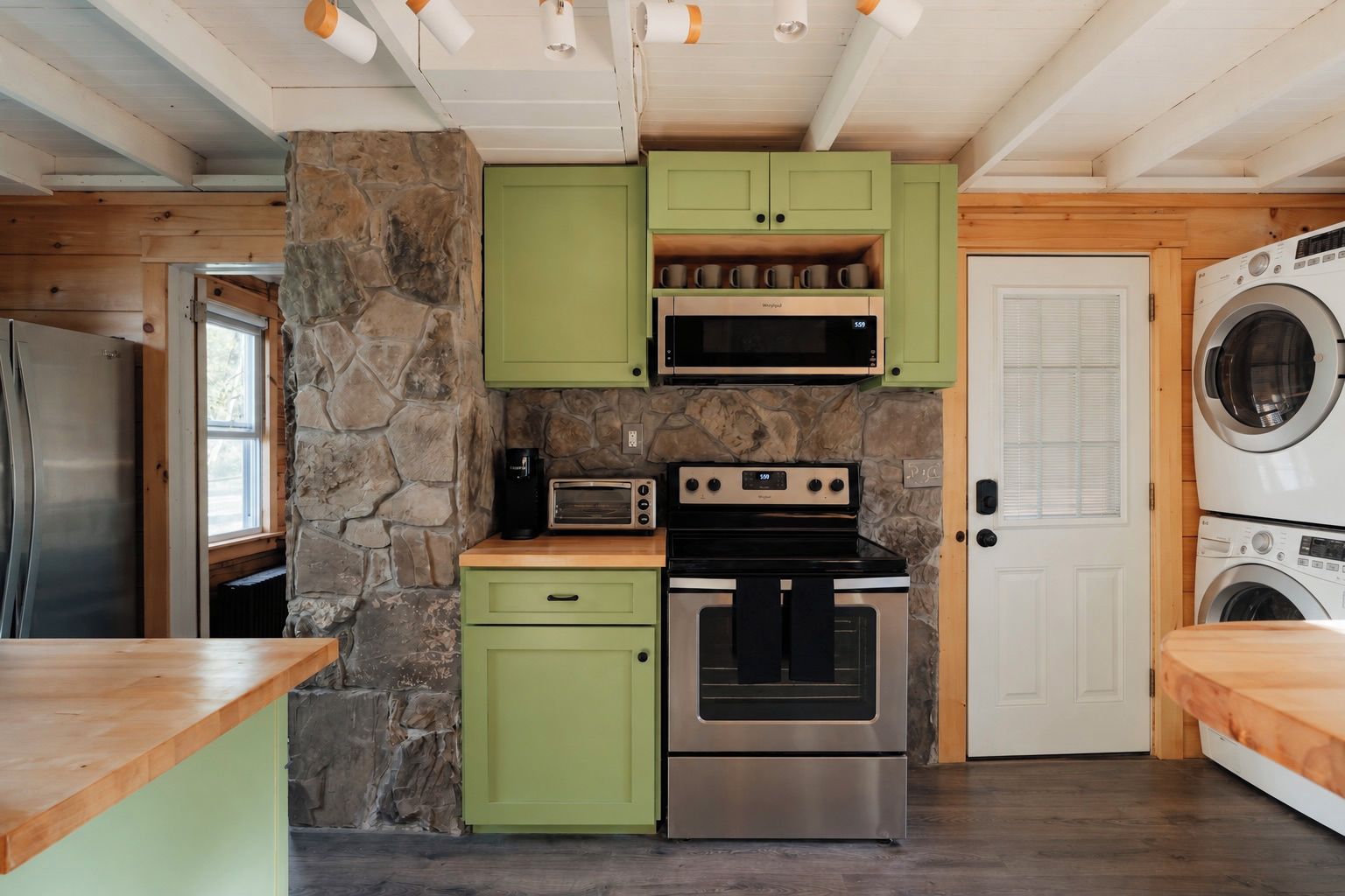 A compact kitchen with a stone accent wall, green cabinets, a stainless steel refrigerator, a microwave, a toaster, an oven, and a washing machine.