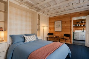 Ein Schlafzimmer mit einem Bett, Nachttischen, einem Wandregal, einem gerahmten Bild und Blick in die Küche.
