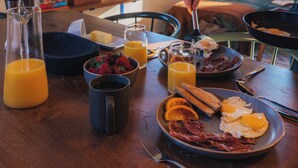 A breakfast table with eggs, bacon, toast, and orange juice. A TV displaying the Netflix logo is mounted on the wall.