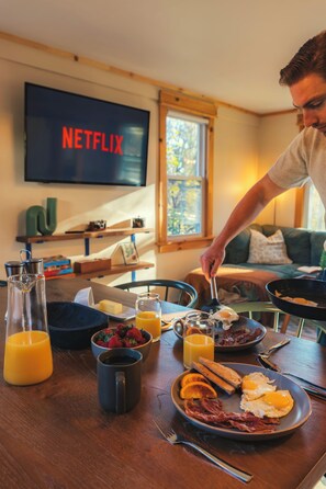 Un desayuno con una televisión mostrando el logo de Netflix, una mesa de comedor con alimentos para el desayuno, y una ventana con luz diurna.