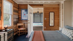 A cozy bedroom with a wooden interior, a large bed, a chair, a desk, and a view of a hot tub through an open door.