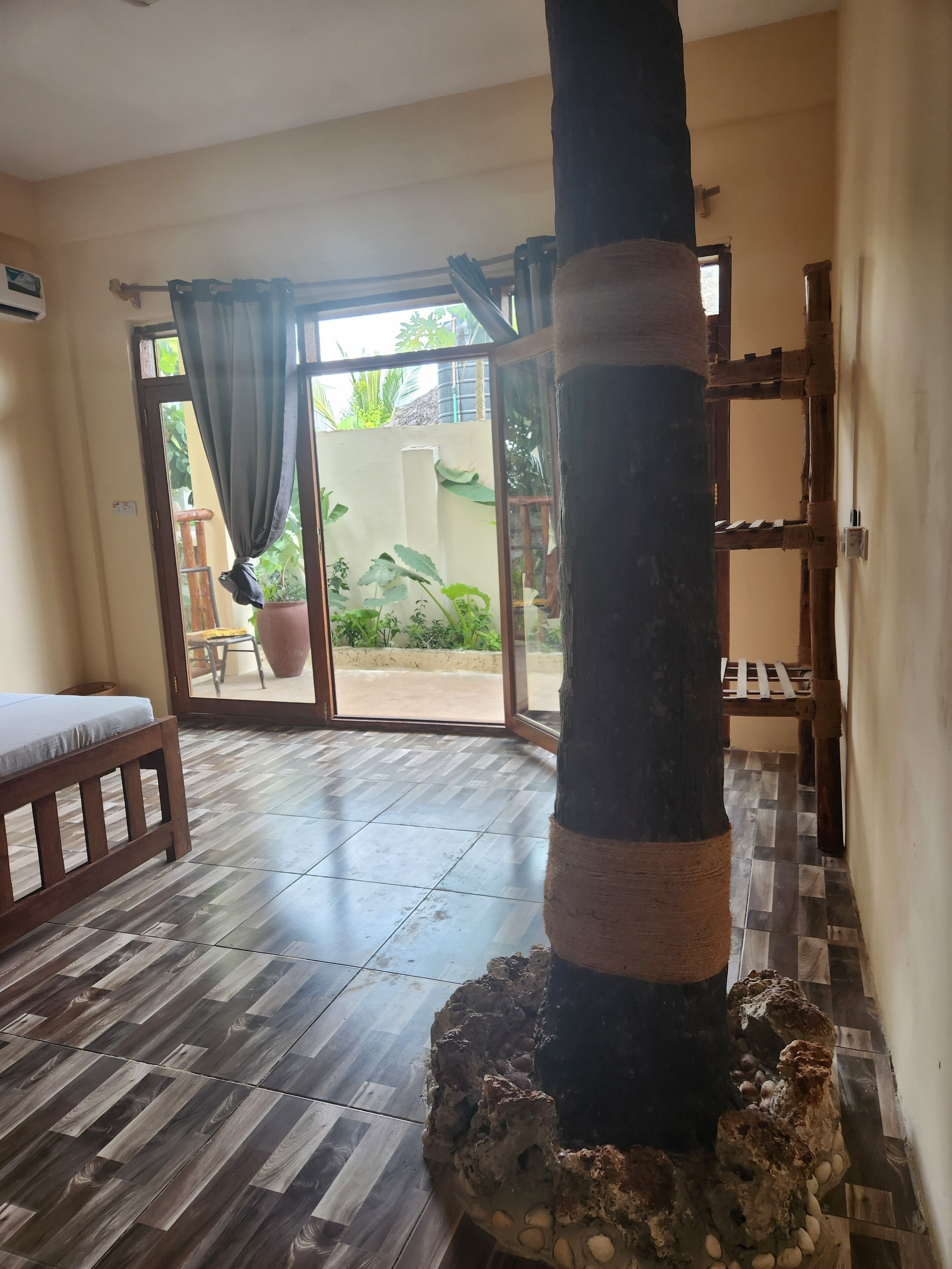 A room with a wooden bench, a pillar with a rope, and a view through a glass door to an outdoor area with plants.