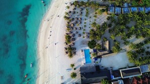 Eine Vogelperspektive zeigt einen Strandort mit Schwimmbad, Palmen und Strandsonnenschirmen.