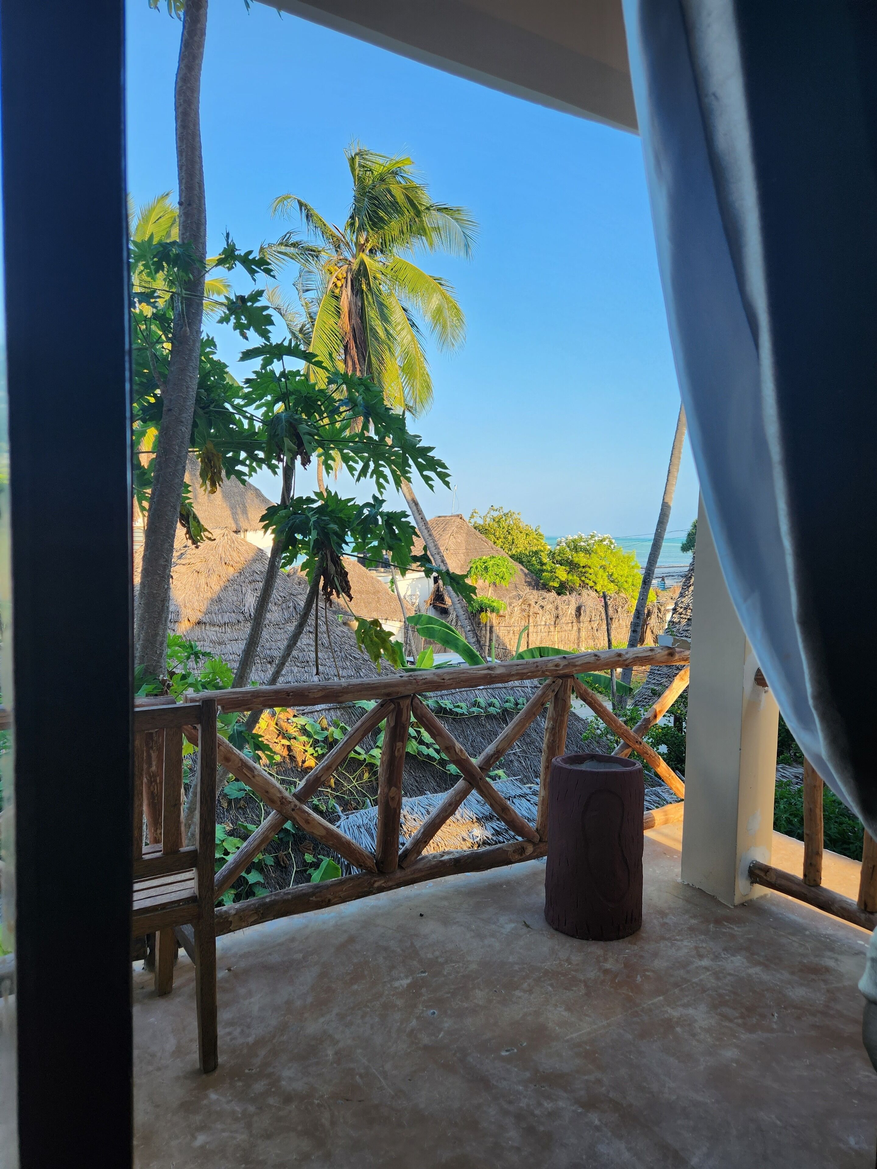 A wooden deck with a thatched roof, overlooking a beach with palm trees and a clear sky.