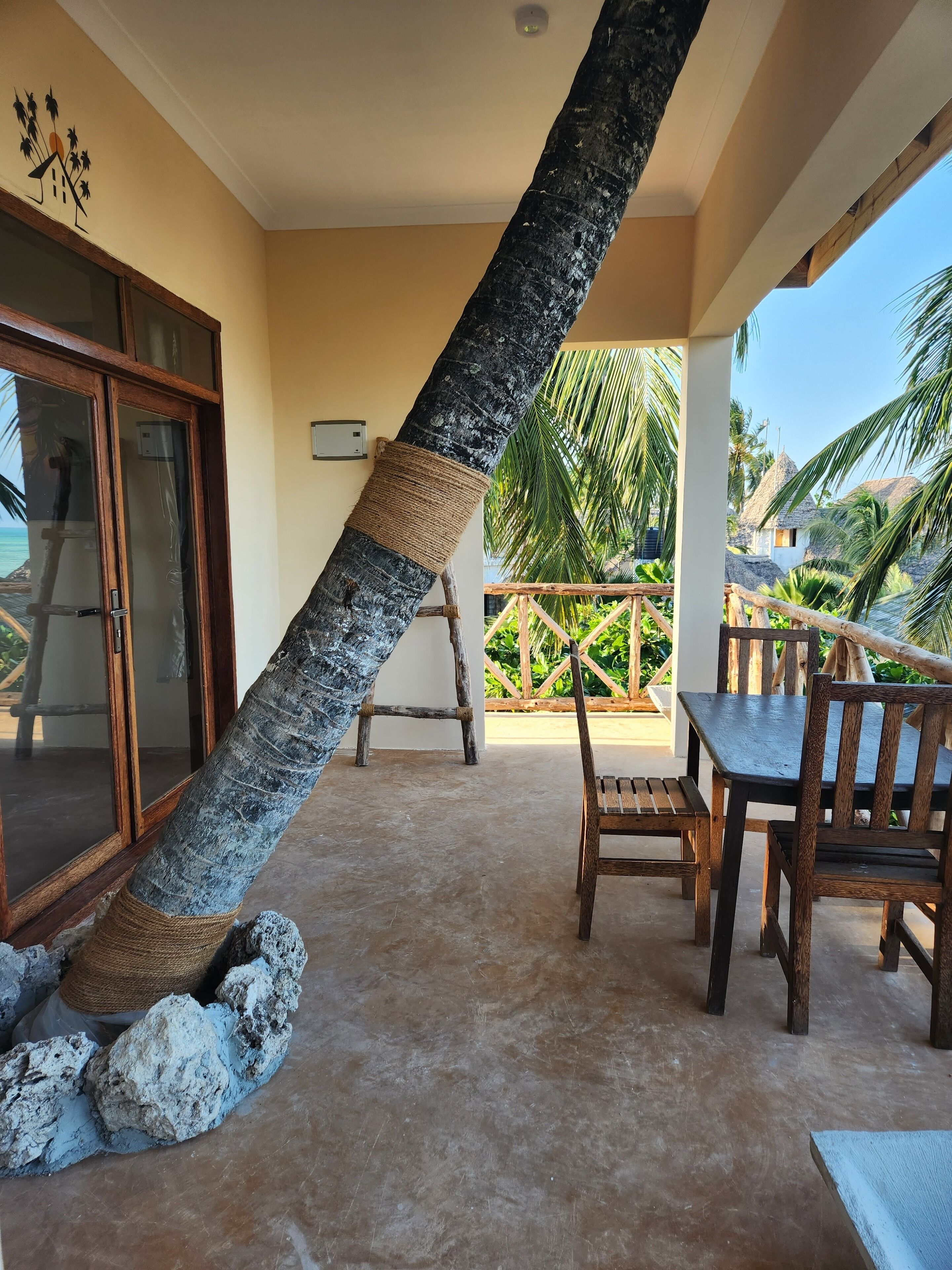 Un espacio al aire libre cubierto con una mesa y sillas de madera, un tronco de árbol grande, y vistas a palmeras y una playa.
