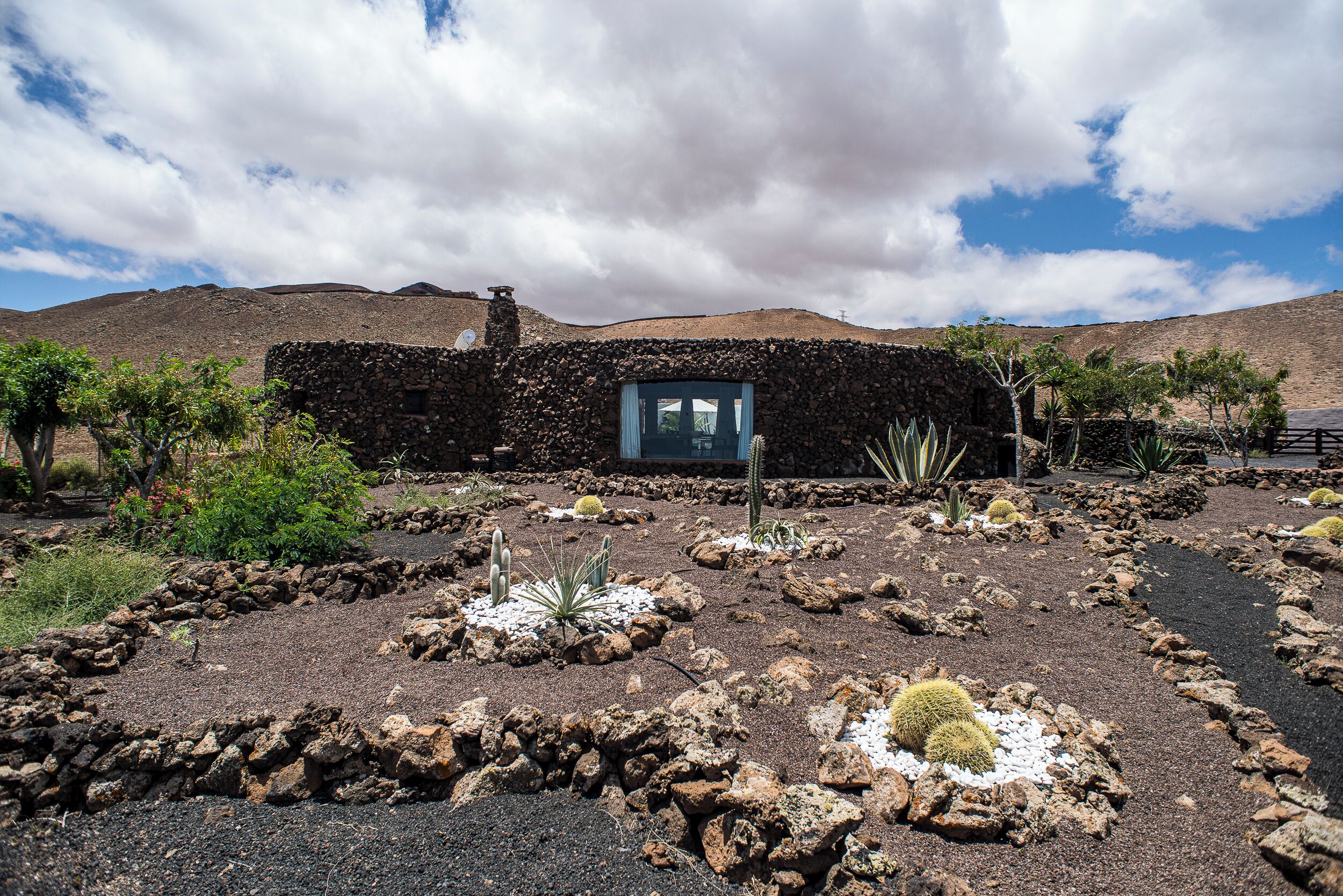 A stone building with a small window, surrounded by a desert garden with cacti and rocks.