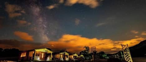 A night scene with a starry sky, a few buildings with lights, and a structure resembling a bridge or tower.