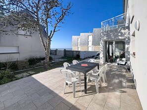 En udendørs terrasse med et bord og stole, et træ og udsigt til havet.