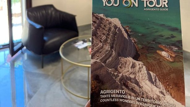 A modern living room with a black leather chair, a floor lamp, and a framed picture on the wall. A brochure titled "You On Tour Agrigento Guide" is prominently displayed. - Calua Luxury Rooms (Agrigento)