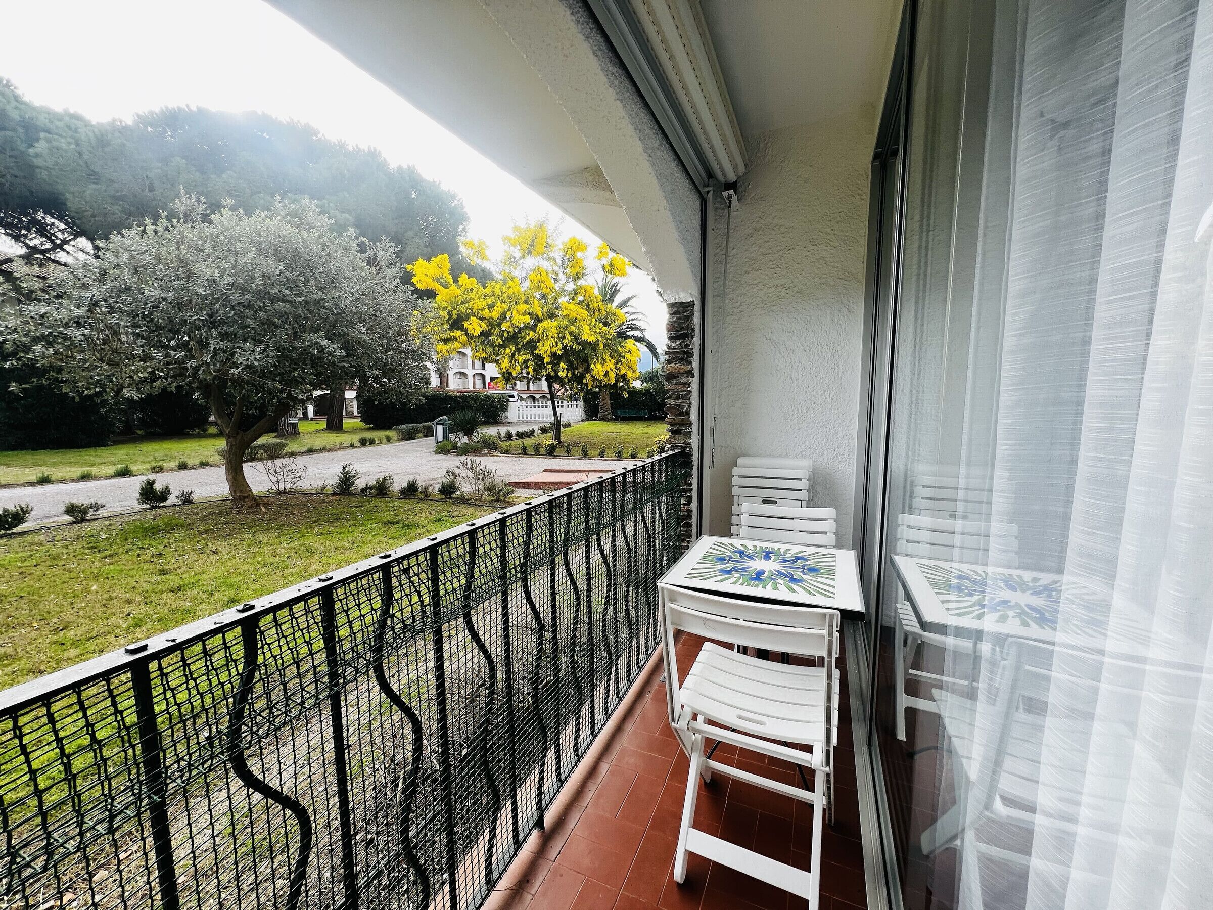 A balcony with a white table and chairs, a black metal railing, and a view of a tree with yellow flowers and a building in the background.