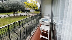 Een balkon met een witte tafel en stoelen, een zwart metalen hekwerk, en uitzicht op een boom met gele bloemen en een gebouw op de achtergrond.