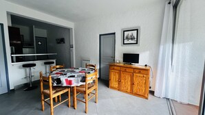 Een knus woongedeelte met een eettafel, stoelen, een kleine keukenhoek, een televisie op een houten kast, en een deur naar een andere kamer.