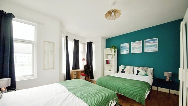 A modern bedroom with two beds, a desk, and a chair. There are three framed pictures on the wall, a plant on a shelf, and a window with curtains. - The Green Garden House -Stunning large house near Shopping centre, Finsbury Park (Greater London)