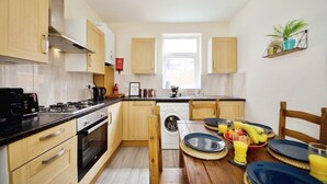 A compact kitchen with a stove, oven, and washing machine, a dining table with plates and glasses, and a window providing natural light. - The Green Garden House -Stunning large house near Shopping centre, Finsbury Park (Greater London)