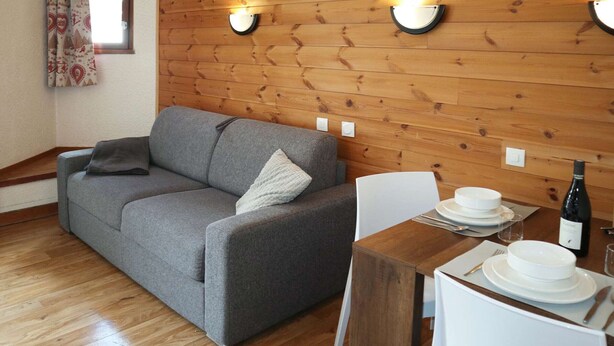 Una stanza accogliente con un divano grigio, un tavolo da pranzo in legno apparecchiato con piatti e una bottiglia di vino, e due luci a parete.