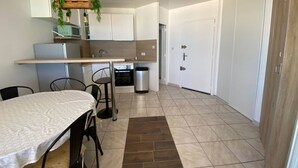 A compact kitchen with white cabinets, a dining table with chairs, a hanging plant, and a trash bin. - Large Studio for 2, Sea View, Private Parking (Le Barcarès)