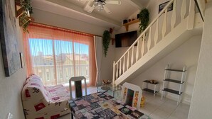 A living room with a sofa, a coffee table covered with a patchwork quilt, a staircase, and a view of the outside through large windows. - Studio Loft 4 people, Pool, Port Barcar ès (Le Barcarès)