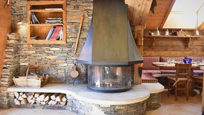 A rustic interior with a stone wall, a wooden bookshelf, a large chimney, and a dining area with wooden furniture.