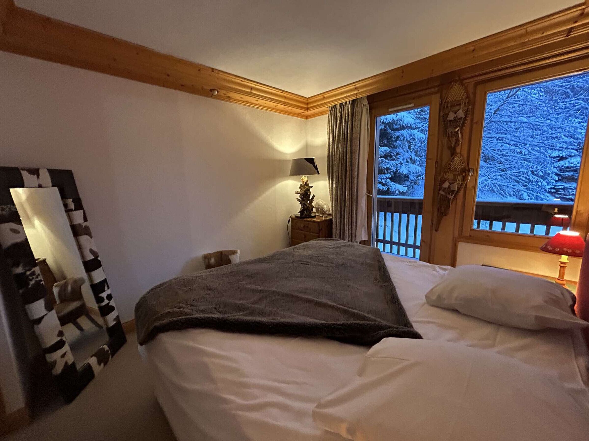 Ein Schlafzimmer mit einem Bett, einem Nachttisch, einer Lampe, einem Fenster mit Blick auf schneebedeckte Bäume und einer Holzbalkendecke.