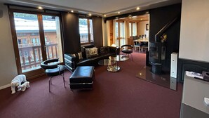 Ein modernes Wohnzimmer mit Sofa, Hocker und Sesseln, einem Kamin und großen Fenstern mit Blick auf Gebäude.