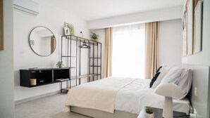A modern bedroom with a bed, a bedside table with a lamp, a black shelving unit, and a large window with curtains.