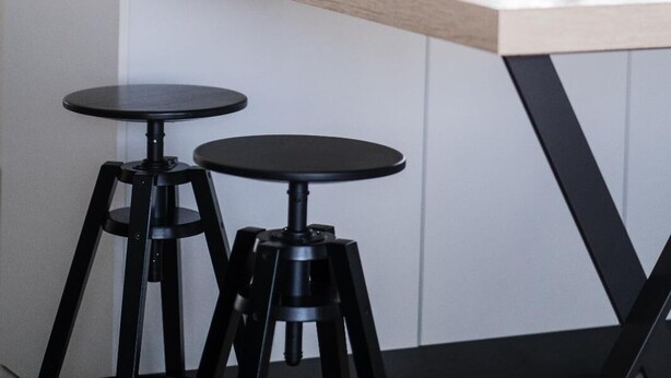 A kitchen with a wooden countertop, two black stools, a potted plant, and a window with blinds.