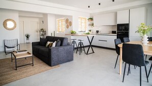 A modern living room with a sofa, coffee table, and bar stools. There is a kitchen area with a dining table and chairs.