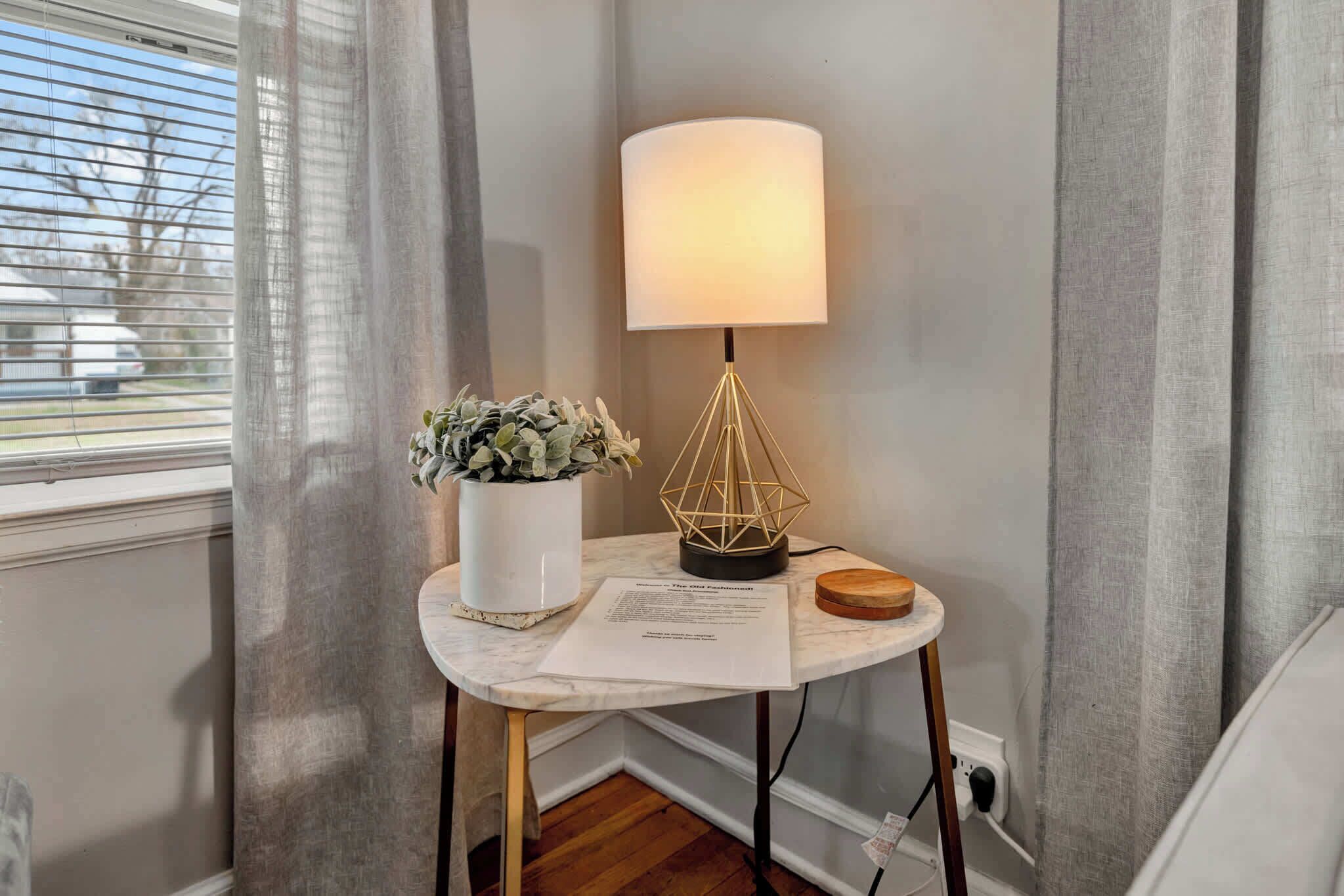 Esquina de una habitación con una mesa de mármol, una lámpara, una planta en maceta y un ventanal con cortinas.
