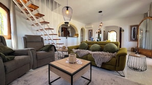 A spacious living room with a staircase, a green sofa, a wooden coffee table, and a unique hanging light fixture.