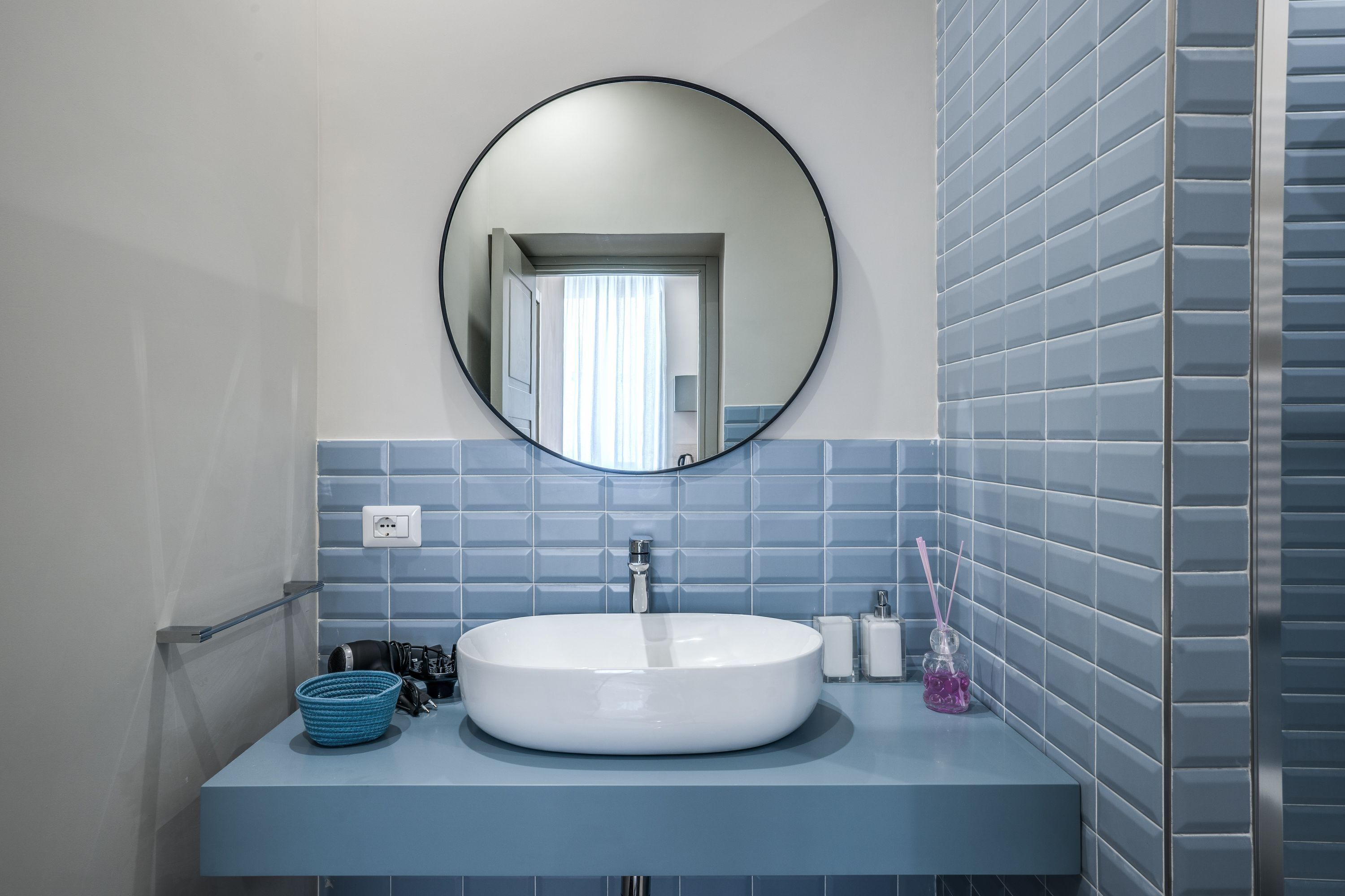 A bathroom with a round mirror, a white sink, and blue tiled walls.