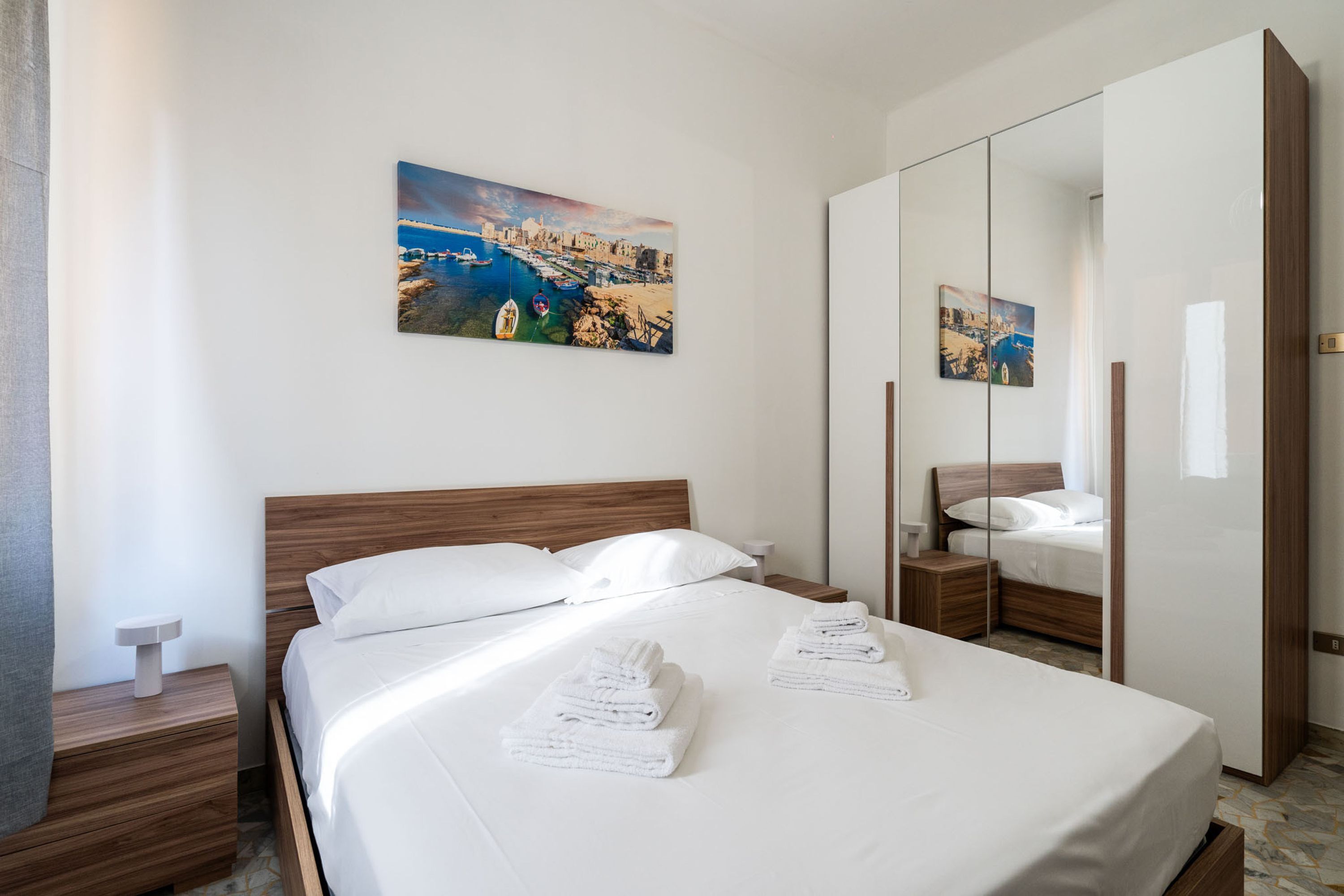 A hotel room with a bed, a nightstand, a mirror, and a painting of a coastal scene.