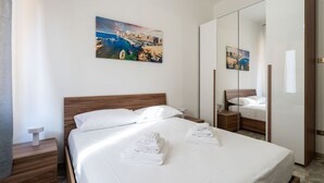 A hotel room with a bed, a nightstand, a mirror, and a painting of a coastal scene.